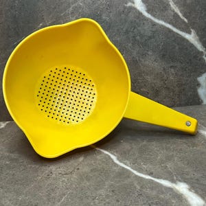 Puede incluir: Un colador de plástico de color amarillo brillante con un cuenco redondo y una base perforada para drenar líquidos. El utensilio de cocina incluye un mango largo con un pequeño orificio circular en el extremo y un pico vertedor moldeado.