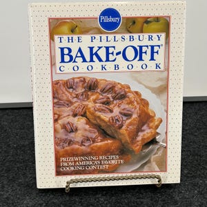 May include: The Pillsbury Bake-Off Cookbook features a white cover with a blue logo and text. The center displays a photograph of a glazed pecan pie on a white plate. The text reads: The Pillsbury Bake-Off Cookbook, Prizewinning Recipes From America's Favorite Cooking Contest.