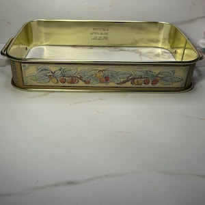 Peut inclure: Un moule de cuisson rectangulaire en métal doré avec des coins arrondis et des poignées latérales. L'extérieur présente une bande décorative avec une illustration détaillée de feuilles vertes et de cerises rouges et orange.