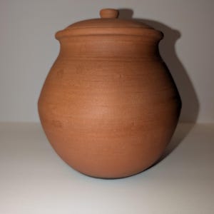 May include: A round, terracotta-colored clay pot with a matching lid. The vessel features a wide, bulbous body that tapers toward a narrow neck and a flared rim. The surface has a matte, earthy texture with subtle horizontal ridges from the crafting process.