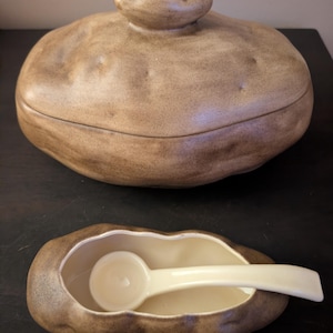 May include: A ceramic lidded container shaped like a potato with a textured brown exterior and a smooth cream-colored interior. A matching smaller open container shaped like a potato includes a cream-colored ceramic spoon resting inside.