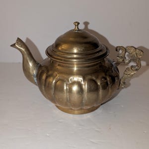May include: A brass teapot with a rounded, fluted body and a matching lid featuring a small knob. The teapot has a curved spout and an ornate, textured handle with intricate detailing. The metal surface shows a warm, aged patina throughout.