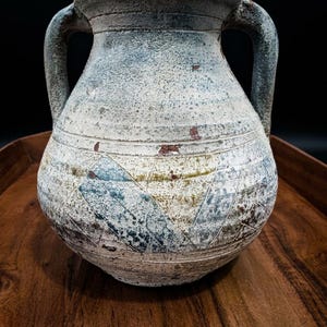 May include: A ceramic vessel with two handles, featuring a weathered, textured finish in shades of white, blue, and brown. The surface displays a geometric pattern of etched lines and patches of color, with a wide mouth and a rounded body shape.