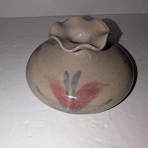 May include: A round, beige ceramic vase with a ruffled, flared rim. The body of the vase features a hand-painted floral motif in muted shades of dusty rose and slate blue, set against a speckled, matte-textured surface finish.