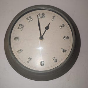 May include: A round wall clock with a dark gray frame and a light beige face. The clock features raised silver-colored numerals from 1 to 12. It has two black hands, with the hour hand pointing toward 1 and the minute hand pointing toward 12.