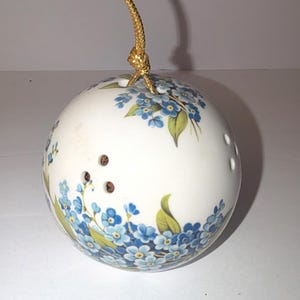 May include: A spherical white ceramic ornament featuring a delicate pattern of blue forget-me-not flowers with green leaves. The ornament has small circular perforations on its surface and is suspended by a thin, metallic gold braided cord.