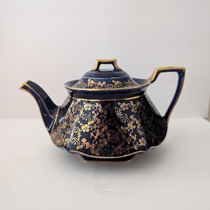 May include: A deep cobalt blue teapot featuring a scalloped body and a lid with a gold floral pattern. The spout, handle, and lid finial are accented with gold trim, and the entire piece displays a repeating intricate gold botanical design.