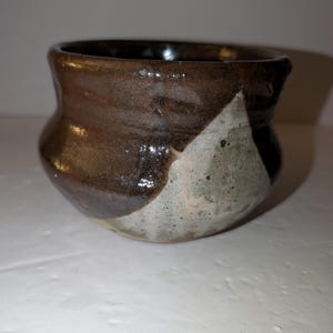 May include: A small, rounded ceramic bowl featuring a two-tone glaze. The upper portion is finished in a glossy, dark chocolate brown, while the lower section displays a speckled, light gray-beige tone with a distinct triangular pattern.