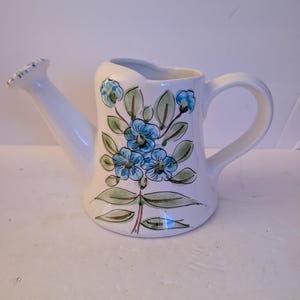 May include: A white ceramic watering can featuring a hand-painted floral design. The decoration consists of blue flowers with green leaves and stems, applied to the front of the vessel. It includes a curved handle and a long, angled spout.