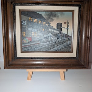 May include: A framed print depicting a steam locomotive at a train station platform. The scene features a dark train with smoke, a building with illuminated windows, and a signal post with the number 143. The artwork is encased in a dark wood frame.