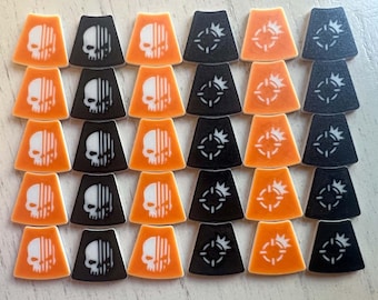 May include: A collection of thirty trapezoidal game tokens arranged in a grid. Fifteen tokens are orange and fifteen are black. Half feature a white skull graphic with vertical lines, while the other half display a white target symbol.