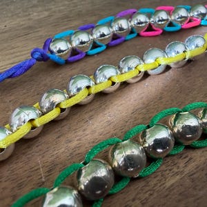 Può includere: Tre braccialetti con perline sferiche color argento lucido infilate su cordoncino di nylon colorato. Un braccialetto usa un cordoncino giallo, un altro un cordoncino verde e il terzo presenta un cordoncino multicolore nei toni viola, blu e rosa.