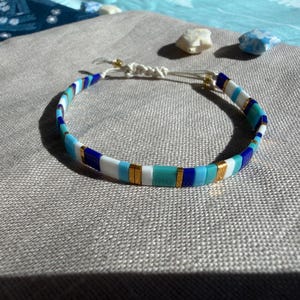 Può includere: Un braccialetto composto da perline rettangolari piatte nei toni del blu reale, turchese, azzurro e bianco, alternate a piccoli distanziatori in metallo dorato. Il braccialetto presenta una chiusura a cordino bianco regolabile con due perline dorate.