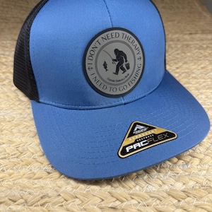 Op de afbeelding: Een blauwe en zwarte honkbalpet met een ronde patch op de voorkant met een afbeelding van een vissend wezen en de tekst I DON'T NEED THERAPY I NEED TO GO FISHING. Een zwart-gouden sticker op de klep bevat de tekst PACFLEX en PACIFIC HEADWEAR.