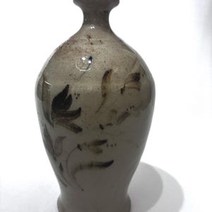 May include: A light beige ceramic vase with a narrow neck and a flared base. The surface features hand-painted dark brown floral motifs and a small blue speck near the neck. The glaze has a speckled texture throughout the body of the vessel.