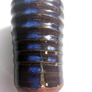May include: A cylindrical ceramic tumbler featuring a ribbed, textured surface with a deep dark brown glaze and a vertical streak of vibrant cobalt blue. The base of the vessel is exposed, unglazed reddish-brown clay, showing a contrast in texture.