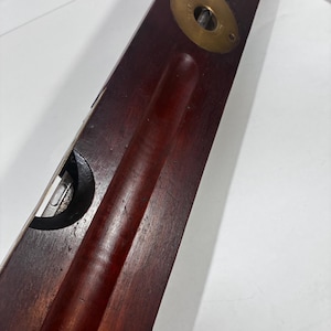 May include: A long, rectangular dark wood spirit level tool featuring a brass circular plate with visible text and a glass vial set into a recessed channel. The tool has a smooth, polished finish and a clear, horizontal bubble level indicator.