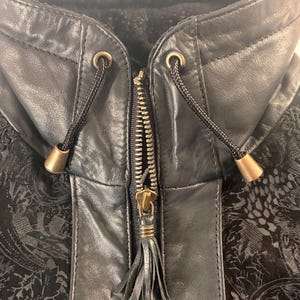 May include: A black jacket featuring a high collar with a drawstring closure, brass-toned toggles, and a central zipper with a leather tassel pull. The garment is constructed from smooth black leather panels combined with a textured black fabric.