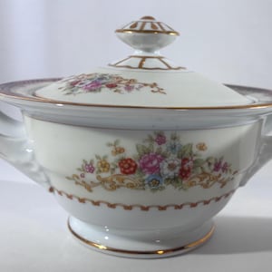 Puede incluir: Un azucarero de porcelana blanca con tapa, decorado con un colorido ramo de flores rosas, rojas y azules. El cuenco tiene dos asas laterales, una base con borde dorado y detalles en oro a lo largo del borde y en el pomo de la tapa.