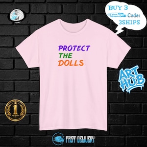 Può includere: Una t-shirt a maniche corte di colore rosa chiaro con la scritta "PROTECT THE DOLLS" stampata in tre colori distinti: "PROTECT" in viola, "THE" in verde e "DOLLS" in arancione, centrata sulla parte anteriore.
