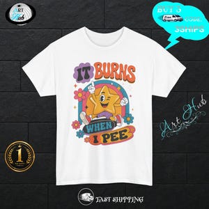 Può includere: Una t-shirt bianca con una grafica colorata di una stella dei cartoni animati che fa l'occhiolino, dotata di braccia e gambe. Il testo recita IT BURNS WHEN I PEE in un carattere retrò. Il design include un arcobaleno, fiori e stelle in arancione, blu e viola.