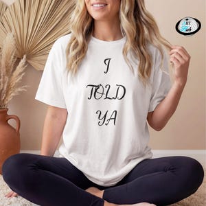 Può includere: Una t-shirt bianca a maniche corte con il testo nero I TOLD YA stampato in un carattere stilizzato sul davanti. Il capo presenta un girocollo e una vestibilità rilassata, offrendo un design semplice e chiaro per un abbigliamento casual.
