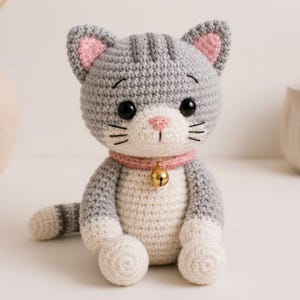 May include: A gray and white crocheted cat plush toy with a pink collar and a small gold bell. The cat features black embroidered whiskers, black plastic eyes, a pink stitched nose, and pink inner ears, with a striped tail and white paws.