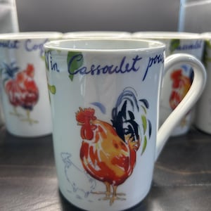 May include: A white ceramic mug featuring a colorful illustration of a rooster with red, orange, and black feathers. The mug displays blue cursive text that reads Vin Cassoulet pour. A smaller outline of a chicken is visible near the rooster.