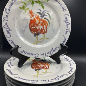 May include: A stack of white ceramic plates featuring a central illustration of a rooster with orange, red, and black feathers. The rim is decorated with green leafy sprigs and the repeating blue cursive text "Coq Au Vin Cassoulet poulet".