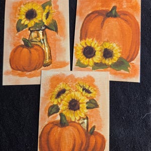 Könnte beinhalten: Drei rechteckige Kunstdrucke mit gemalten Herbstszenen. Jeder Druck zeigt leuchtend gelbe Sonnenblumen, orangefarbene Kürbisse und Glasvasen vor einem warmen, strukturierten orangefarbenen Hintergrund, die ein herbstliches Erntethema darstellen.