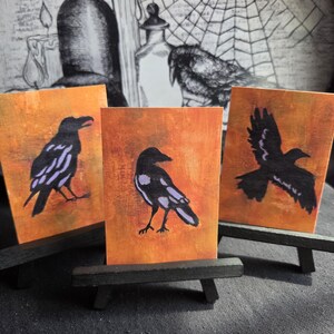 Può includere: Tre cartoline artistiche rettangolari con illustrazioni di corvi neri con accenti viola sulle ali su uno sfondo arancione strutturato. Ogni cartolina è esposta su un piccolo cavalletto di legno nero, mostrando diverse pose degli uccelli.