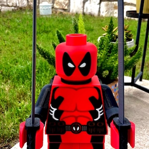 May include: A large red and black plastic figure resembling a comic book character with a round head, black eye patches, and a muscular torso design. The figure holds two long, thin black swords in its hands and has a gray plastic weapon at its feet.
