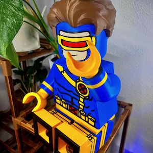 May include: A large, 3D-printed plastic figure of the character Cyclops from X-Men. The figure features a blue suit with yellow accents, a yellow utility belt with red X-logos, brown hair, and a signature red visor over the eyes with yellow frames.