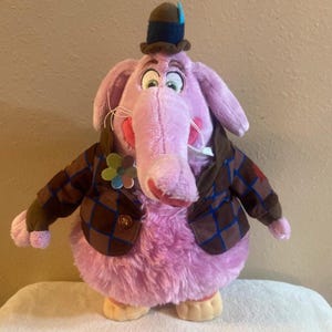 May include: A plush toy character with light purple fur, a long trunk, and large ears. It wears a brown and blue plaid jacket with a flower pin, a brown hat with a blue feather, and has large white eyes with black pupils and light green irises.