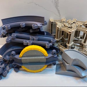 Puede incluir: Un conjunto de componentes de vías de tren de juguete de plástico que incluye piezas curvas y rectas de color gris, una plataforma giratoria gris y amarilla, y estructuras de soporte beige para construir diseños de vías elevadas.