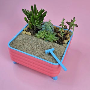 May include: A rectangular pink planter with a light blue rim and four small light blue feet. The planter contains sand, small pebbles, and several green succulents. A small light blue rake rests on the sand surface inside the container.