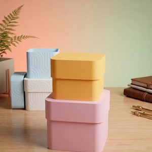 May include: A stack of two square boxes in yellow and pink, positioned in front of two additional square boxes in light blue and white, each featuring a raised, curved line pattern on the exterior surfaces.