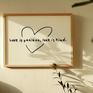 Peut inclure: Une décoration murale rectangulaire dans un cadre en bois clair, présentant un fond blanc avec le contour d'un cœur noir dessiné à la main. Le texte noir "Love is patient, love is kind." est inscrit au centre dans une police manuscrite.