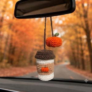 Peut inclure: Un accessoire de voiture en crochet suspendu au rétroviseur, composé d'une tasse à café blanche avec un couvercle marron et une manchette beige ornée d'une petite citrouille orange, accompagnée d'une petite citrouille orange à tige verte.