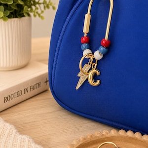 May include: A gold-toned U-shaped carabiner keychain attached to a blue bag. The keychain features red, blue, and white beads, along with two dangling gold charms: a lightning bolt with small clear stones and a letter C in a metallic finish.