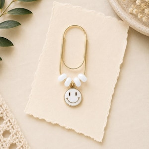 May include: A gold-toned metal paperclip featuring four small white beads threaded onto the bottom loop, with a circular white enamel charm dangling from the center, displaying a black line-drawn smiley face with two eyes and a curved mouth.