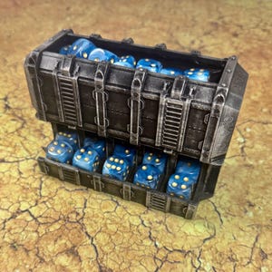 May include: A two-tiered rectangular storage container with a weathered dark metallic finish, filled with multiple translucent blue six-sided dice featuring gold pips. The container has a reinforced industrial design with vertical support beams.