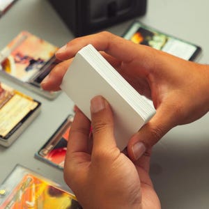 Puede incluir: Un mazo de cartas de juego blancas en blanco sostenido en la mano, rodeado por varias otras cartas coleccionables individuales con ilustraciones coloridas y texto, dispuestas sobre una superficie plana.