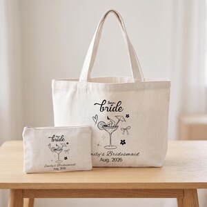 May include: A cream-coloured canvas tote bag and a matching zippered pouch, both featuring a black graphic of a cocktail glass with legs, a bow, a heart, and the text "team bride", "Emily's Bridesmaid", and "Aug. 2026" printed on the front.