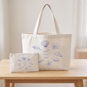 May include: A cream-coloured canvas tote bag and a matching smaller zippered pouch, both featuring a blue printed design of various seashells and starfish arranged in a circle around the text salt sand soul in a cursive script font.