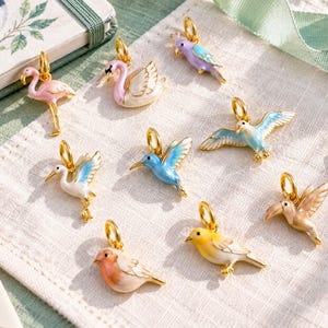 Può includere: Una collezione di nove ciondoli in metallo a forma di vari uccelli, tra cui un fenicottero rosa, un cigno bianco, un pappagallo viola, un colibrì blu, una cicogna bianca, un gabbiano blu, un uccellino giallo e un tucano beige, con anello.