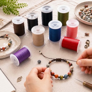 Könnte beinhalten: Eine Sammlung bunter Garnrollen in Braun, Schwarz, Beige, Blau, Rot, Grün und Violett, zusammen mit zwei Perlenarmbändern, einer kleinen Schale mit losen Steinperlen, einem Metallblatt-Anhänger, einer Schere und einem handbeschriebenen Notizbuch.