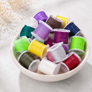 May include: A collection of small plastic spools wrapped with colorful cord in shades of purple, yellow, brown, dark blue, green, lime green, silver, white, and red, all contained within a shallow, round, off-white ceramic bowl.