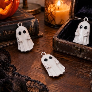 May include: Three white ghost-shaped metal charms with black circular eyes. Each ghost figure features two small arms raised, with each hand displaying a middle finger gesture. A small silver metal loop is attached to the top of each ghost charm.