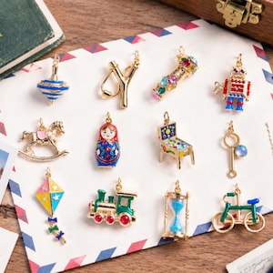 May include: A collection of twelve gold-tone metal charms featuring colourful enamel and rhinestone details. The charms include a spinning top, slingshot, kaleidoscope, robot, rocking horse, nesting doll, pinball machine, bubble wand, kite, train, hourglass, and bicycle.